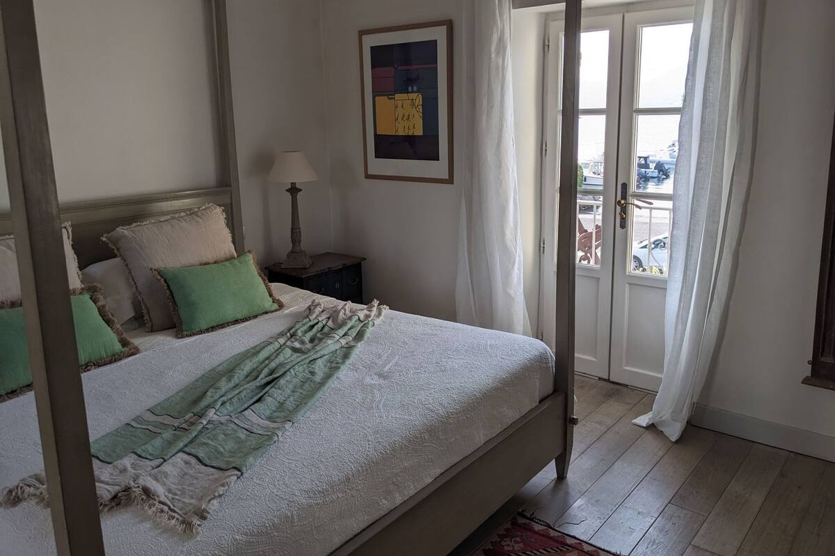 A four-poster bed with a white bedspread and green pillows, a bedside table with a lamp, and a view of the outside through a door.