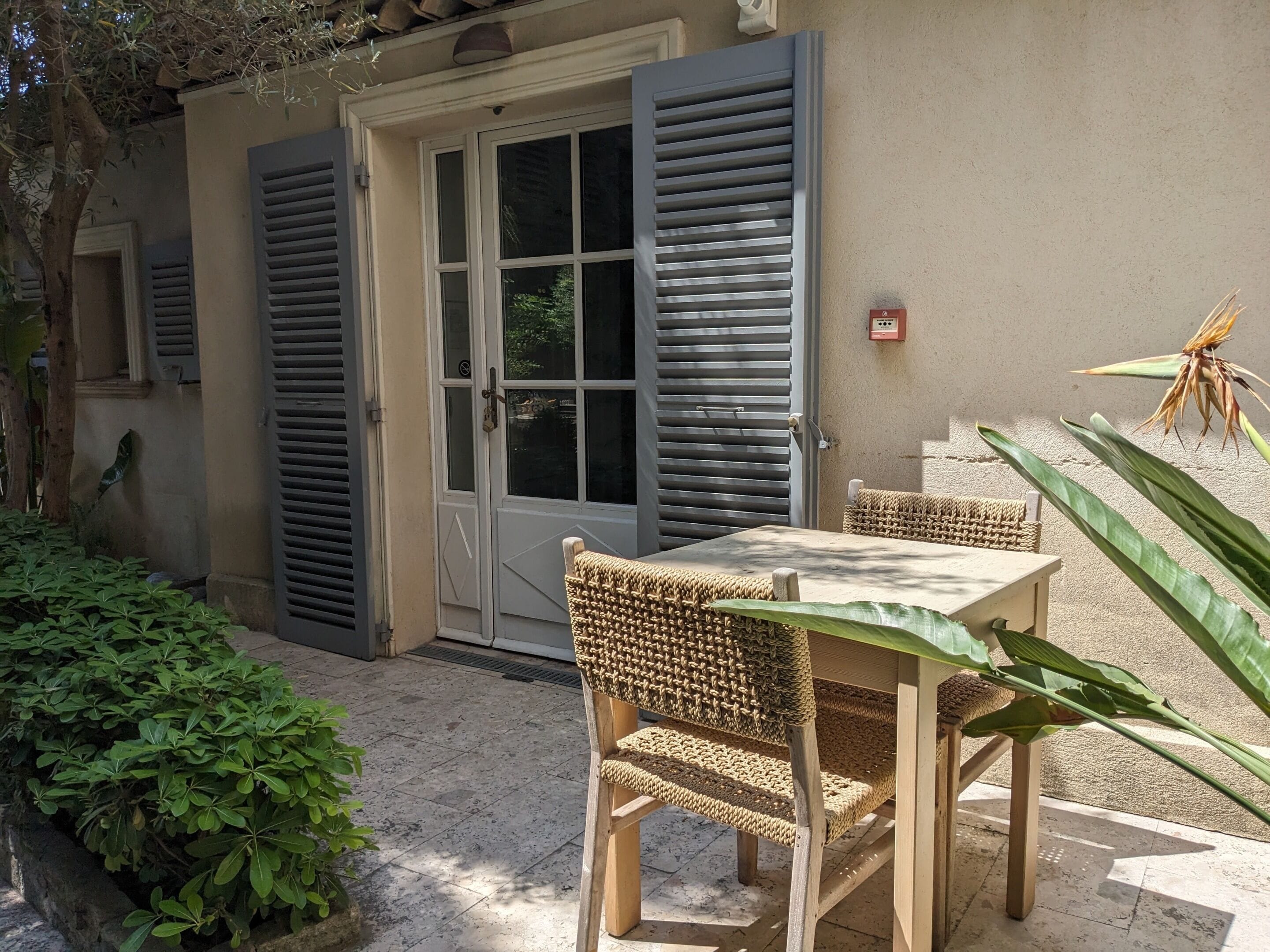 Une terrasse avec une table et des chaises, une porte avec des volets et un petit jardin.