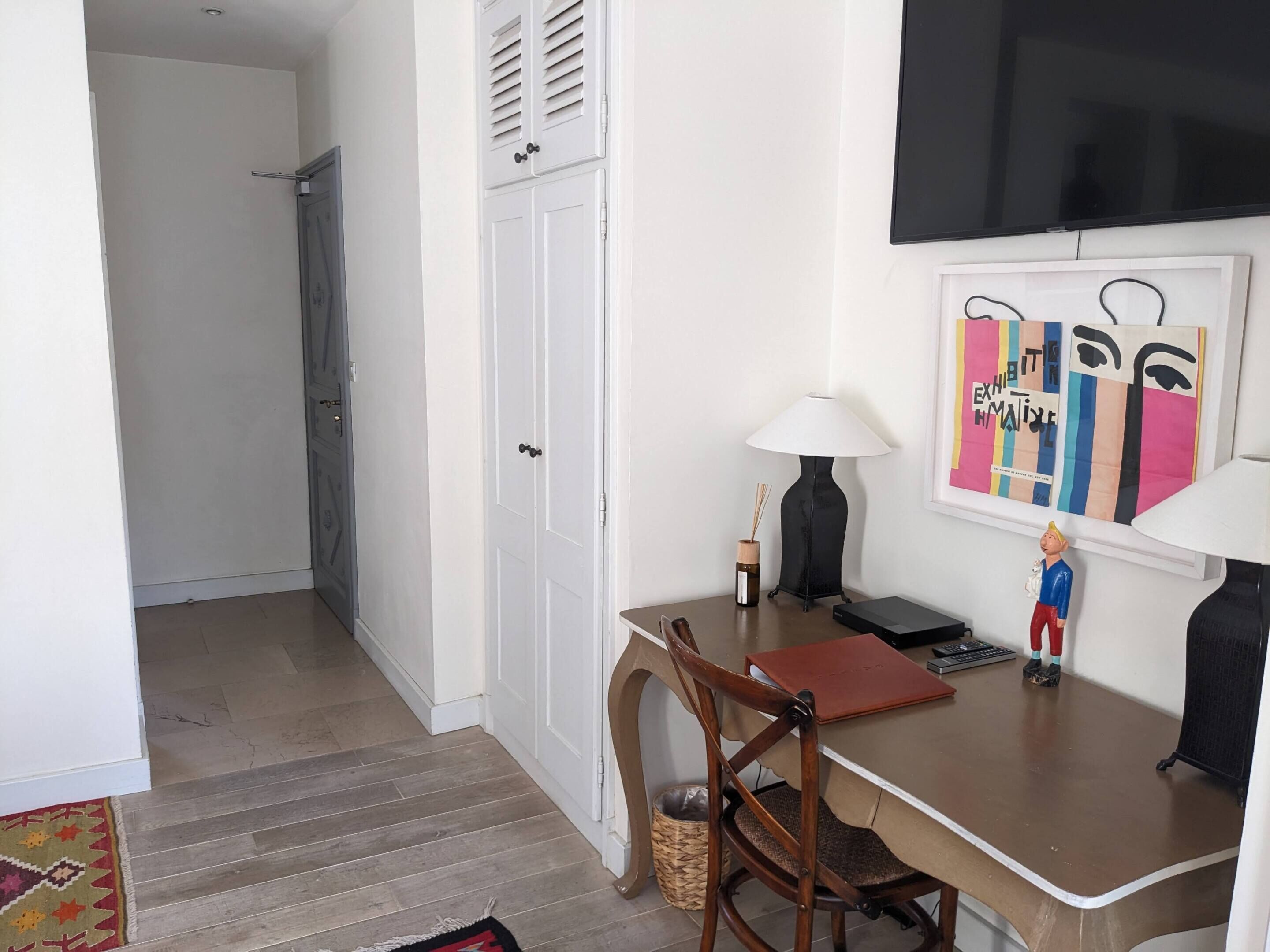Ein kleiner, ordentlicher Raum mit Schreibtisch, Stuhl, Lampe und einem an der Wand befestigten Fernseher.