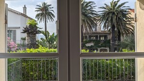 A view through a window with a balcony, featuring a white building with a tiled roof, palm trees, and a stone planter with green plants. - Pastis Hotel Saint-Tropez (Saint-Tropez)