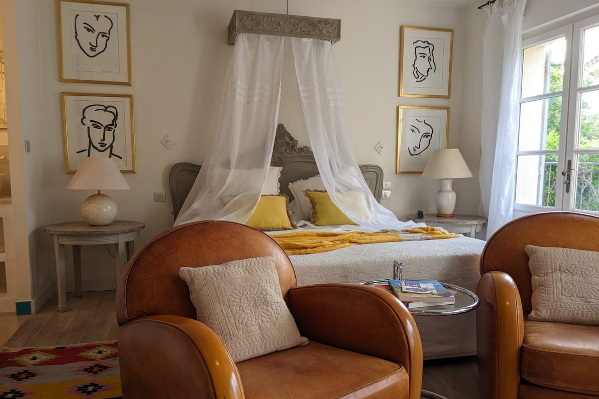 A bedroom with a four-poster bed, two bedside tables with lamps, a small round table, and two leather armchairs.