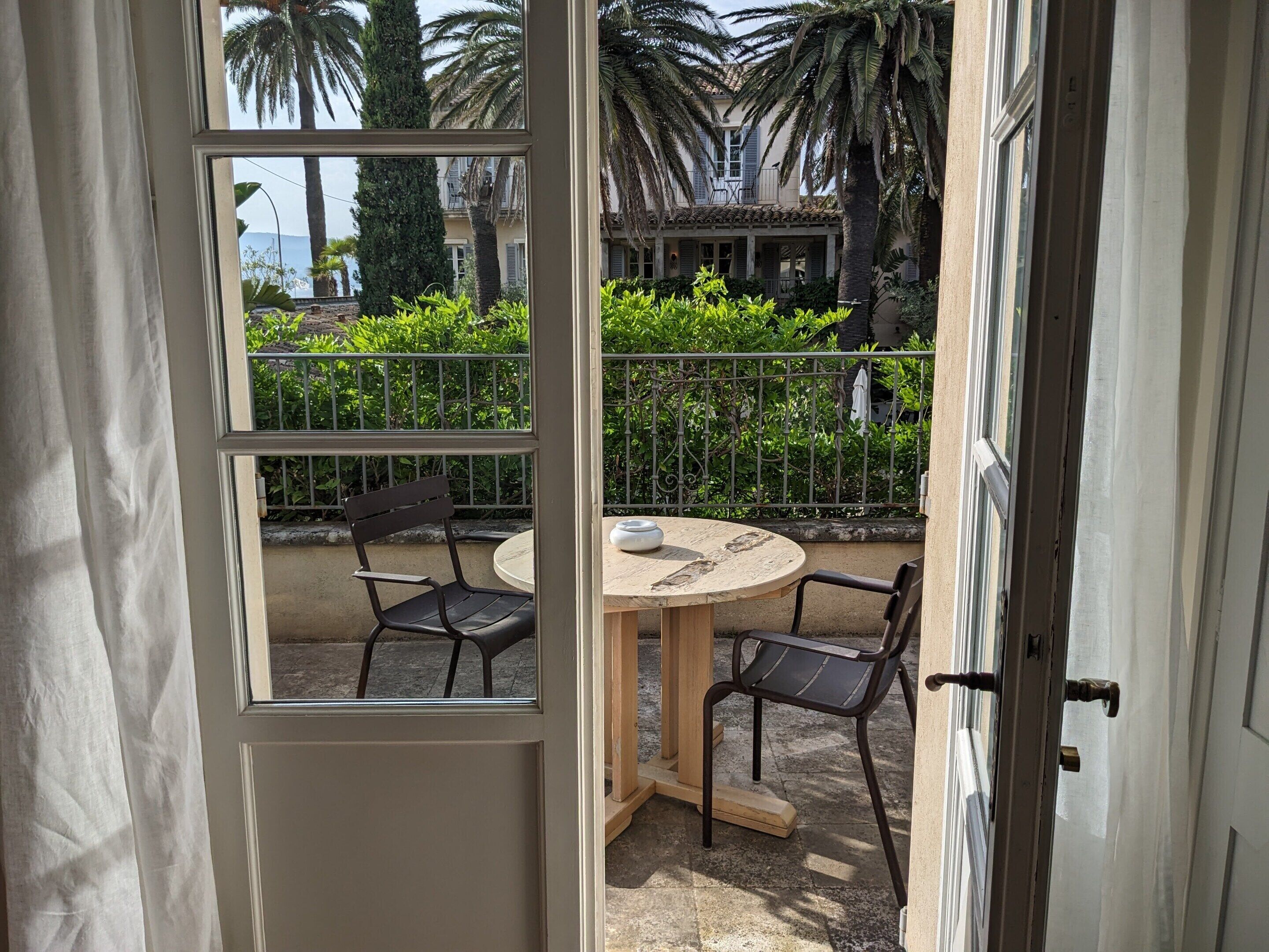 Un balcón con una mesa redonda y dos sillas, con vista a un jardín con palmeras y un edificio al fondo.