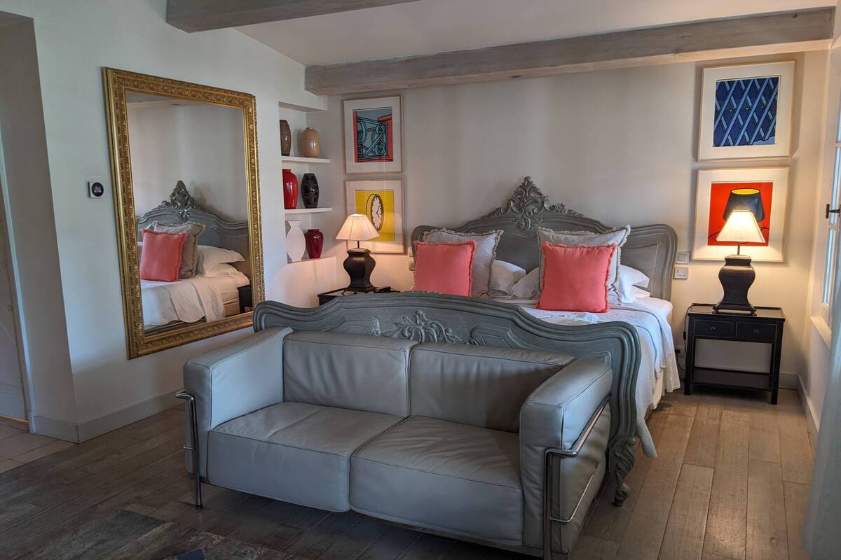 A bedroom with a large bed, a grey sofa, a bedside table with a lamp, and a mirror on the wall.