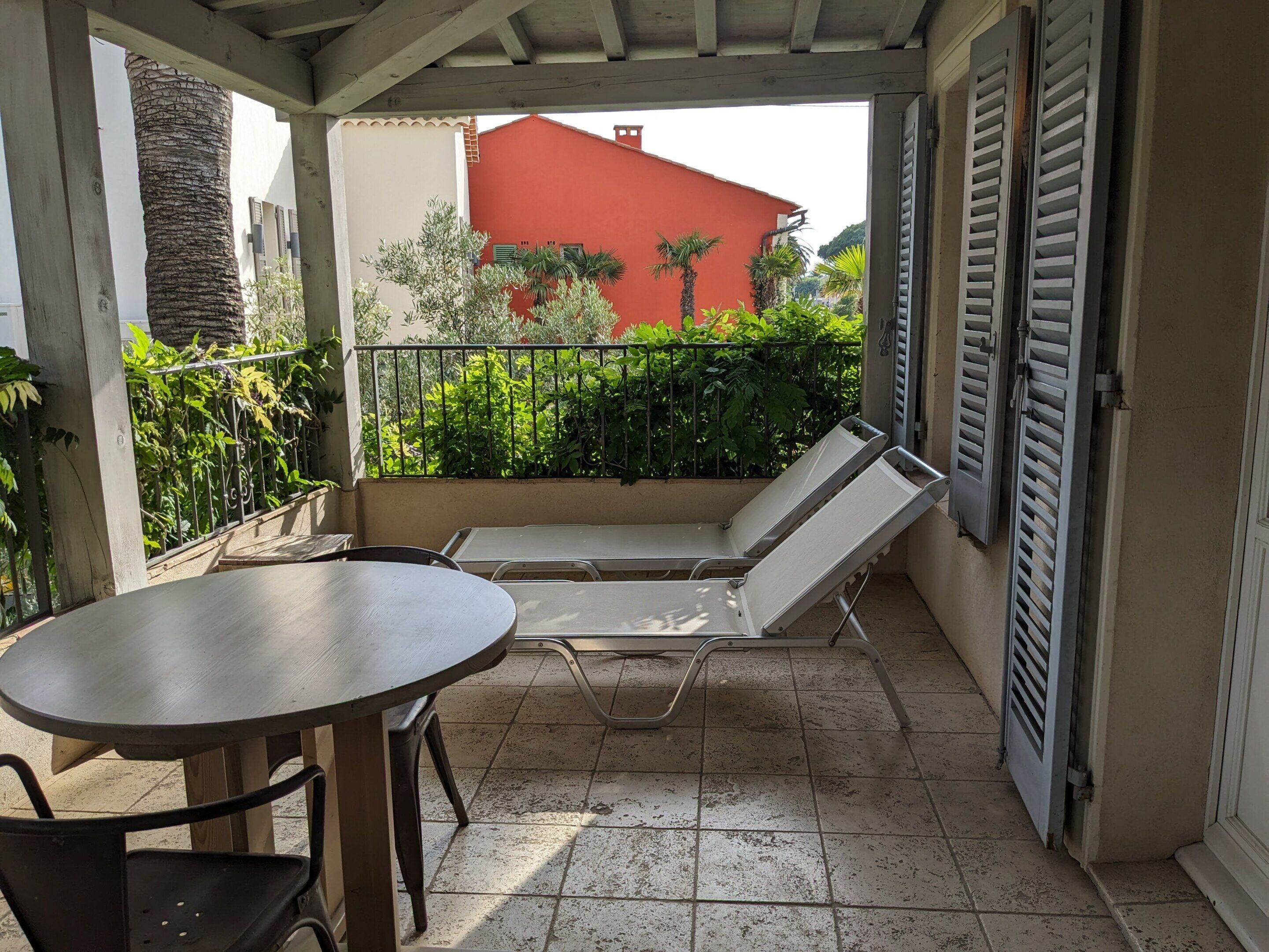 Ein Balkon mit Tisch und zwei Lounge-Stühlen, umgeben von Grünpflanzen und einem roten Gebäude im Hintergrund.