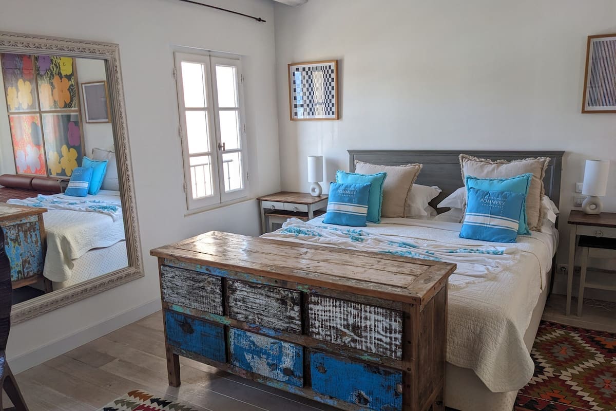A bedroom with a large mirror, a wooden chest of drawers, a bed with blue and beige bedding, and a patterned rug.