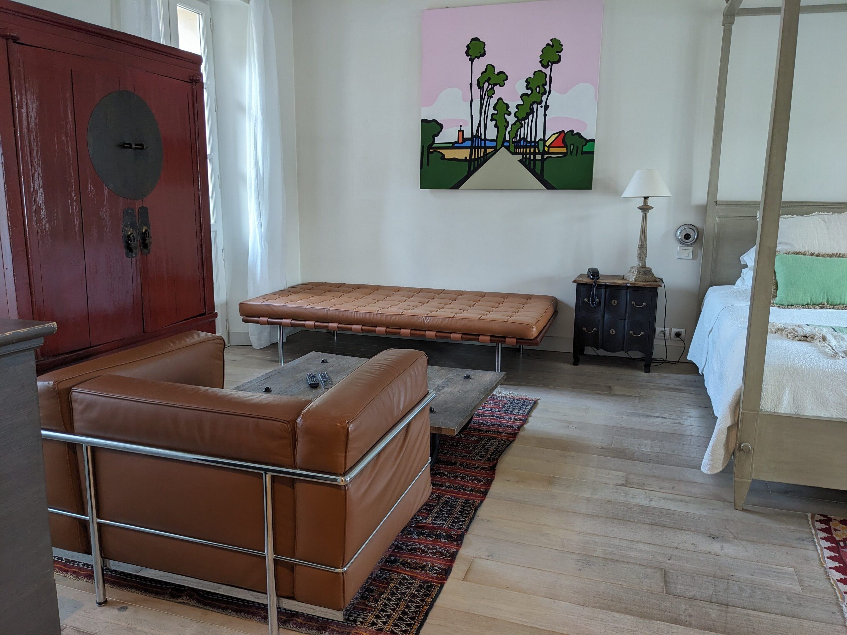 A room with a brown leather sofa, a bed with white bedding, a small wooden nightstand, a lamp, and a painting on the wall.