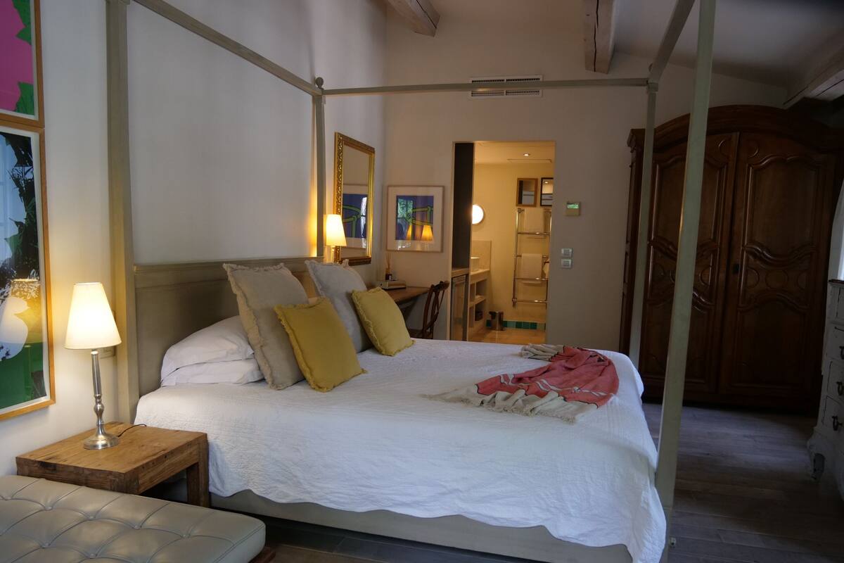 A four-poster bed with a white bedspread and pillows, a bedside lamp, a wooden nightstand, and a bench with a cushioned seat.