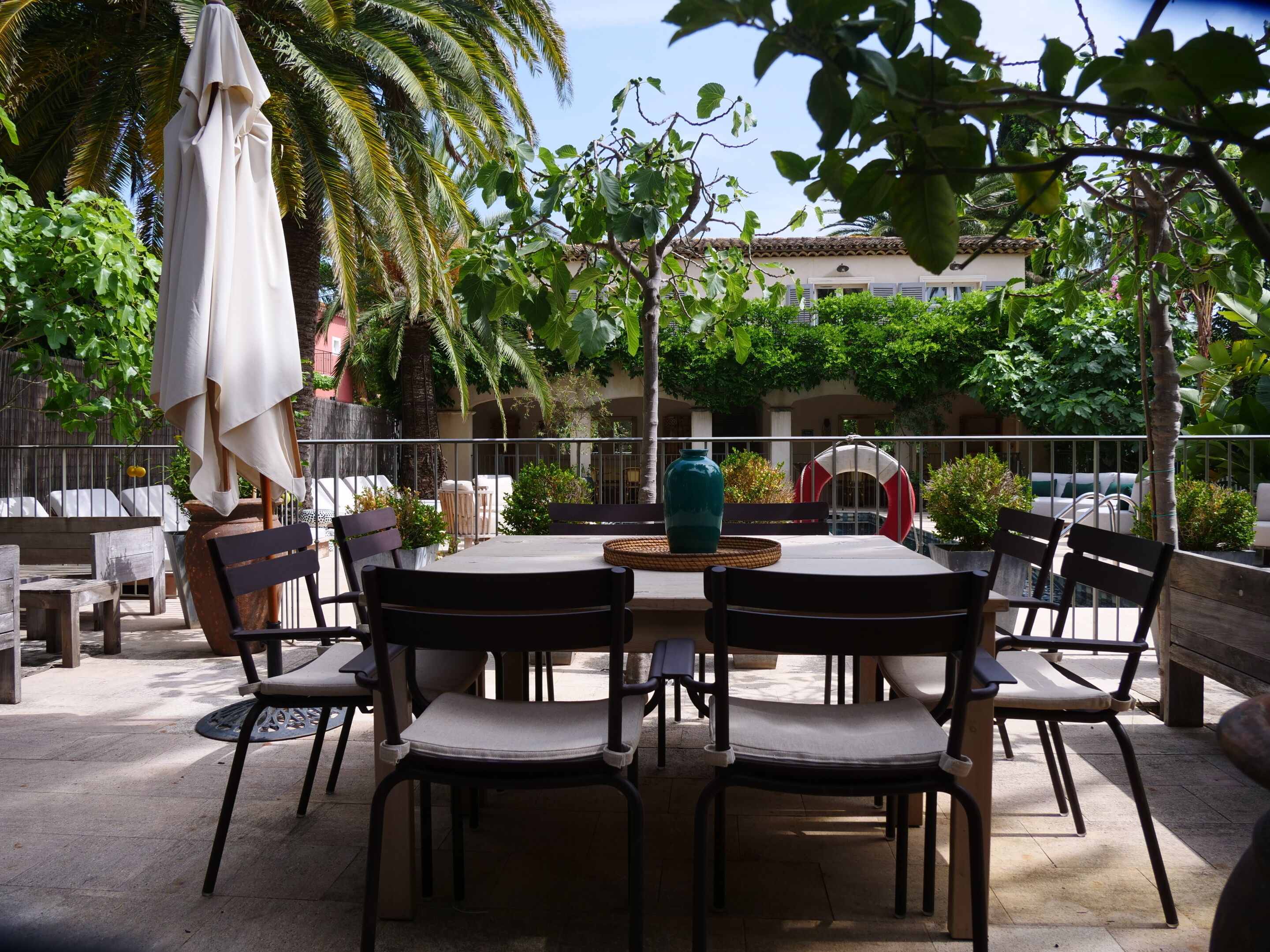 An outdoor patio with a round table and chairs, surrounded by lush greenery and a palm tree.
