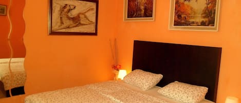 Een slaapkamer met een groot bed, twee ingelijste schilderijen en een nachtkastje met een lamp.
