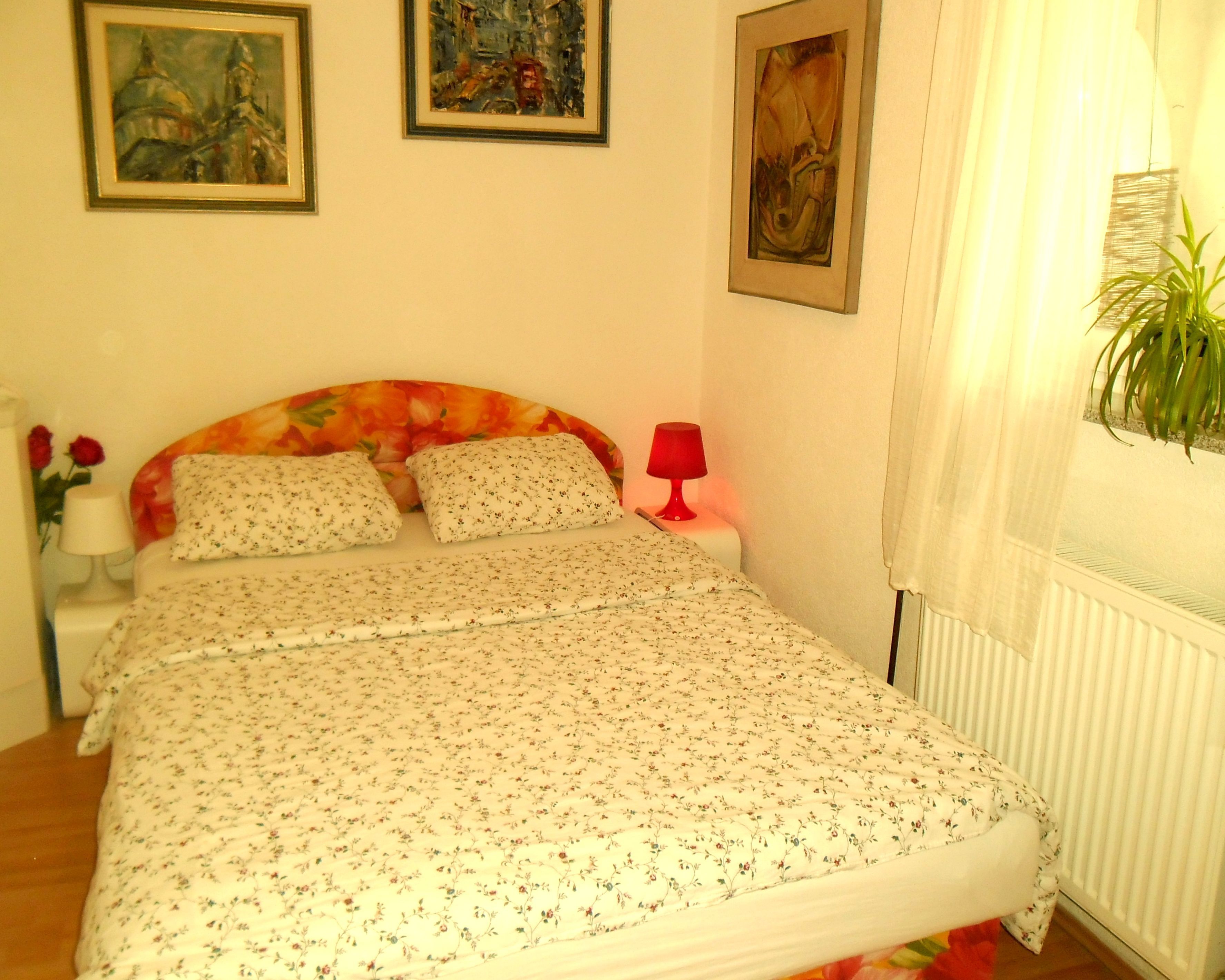 Ein Schlafzimmer mit einem blumigen Bettbezug, zwei Kissen, einer roten Nachttischlampe und einem Blumentopf am Fenster.