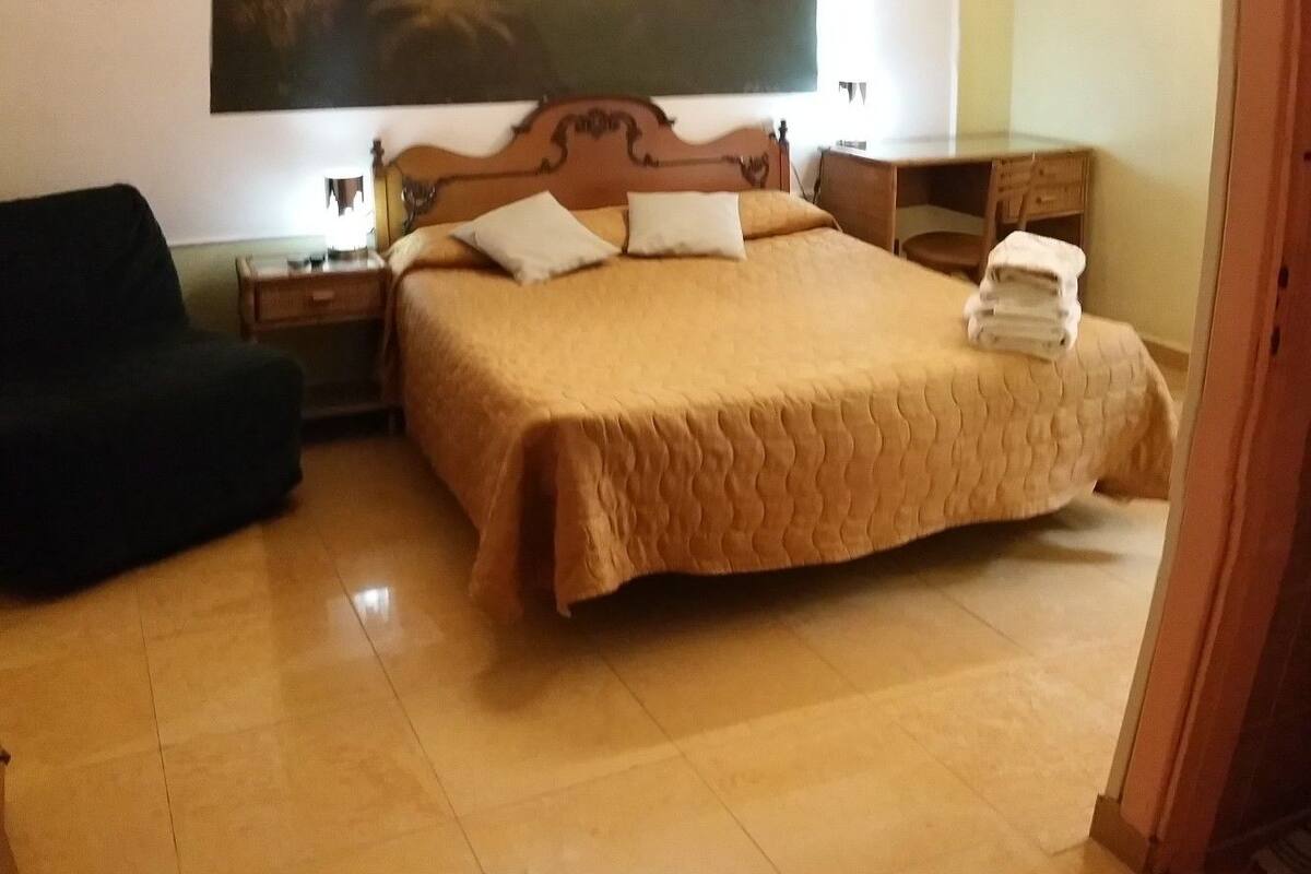 A bedroom with a bed, a desk, a chair, a nightstand, a ceiling fan, and a painting on the wall.