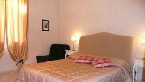 A neatly made bed with a beige quilt, two pillows, a black chair, and a bedside table with a lamp. - Walter Guest House (Rome)