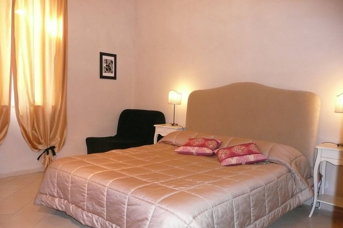 A neatly made bed with a beige quilt, two pillows, a black chair, and a bedside table with a lamp.