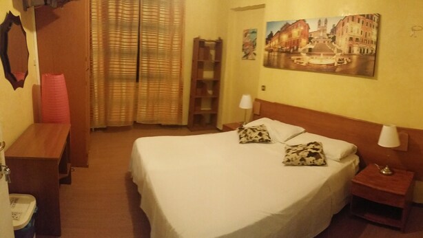A hotel room with a bed, a nightstand, a lamp, a red trash bin, a wooden bench, a mirror, a window with blinds, and a painting on the wall. - Walter Rooms (Rome)