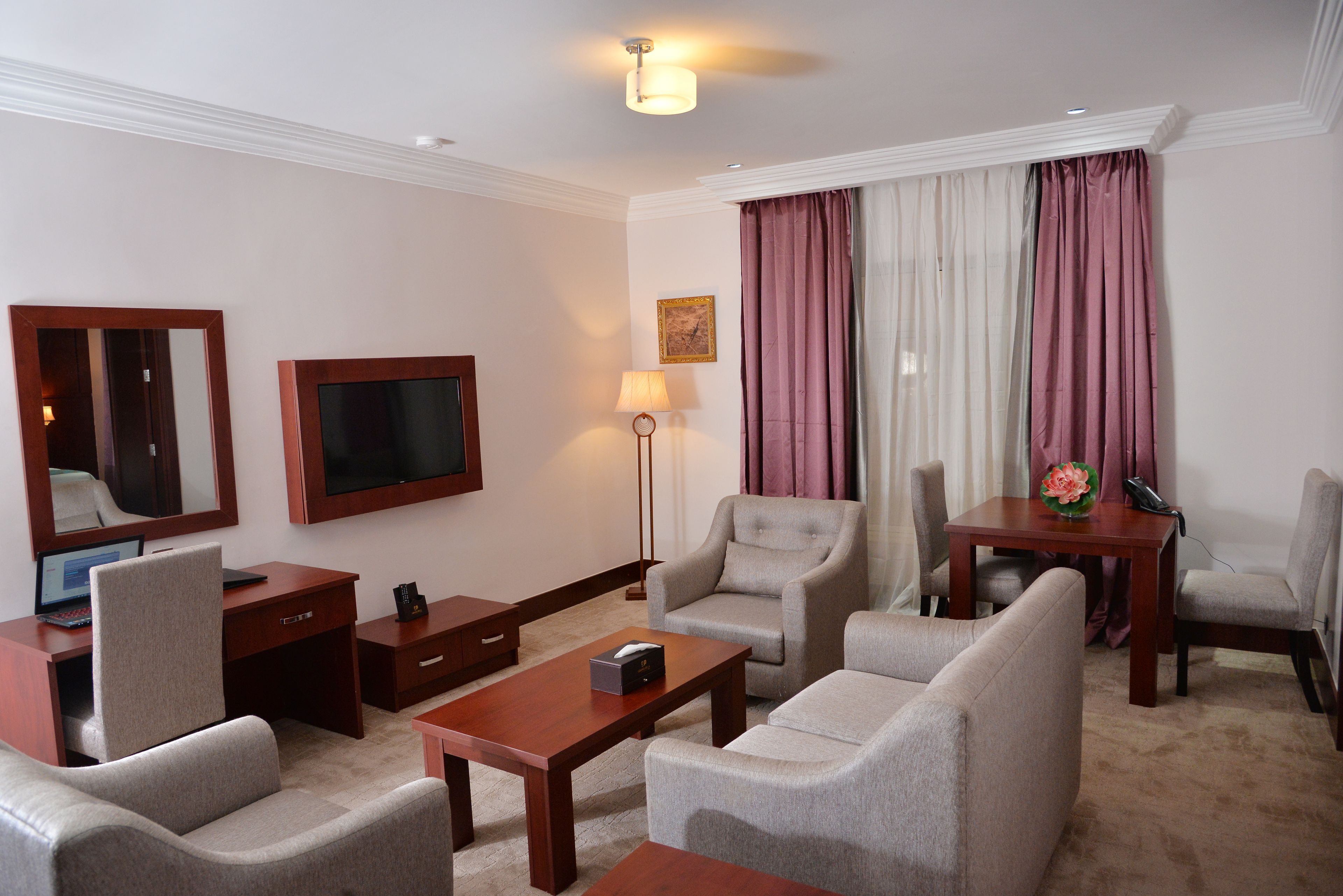 Quarto de hotel com escrivaninha, duas poltronas, um sofá, televisão, abajur e mesa com um vaso de flores.