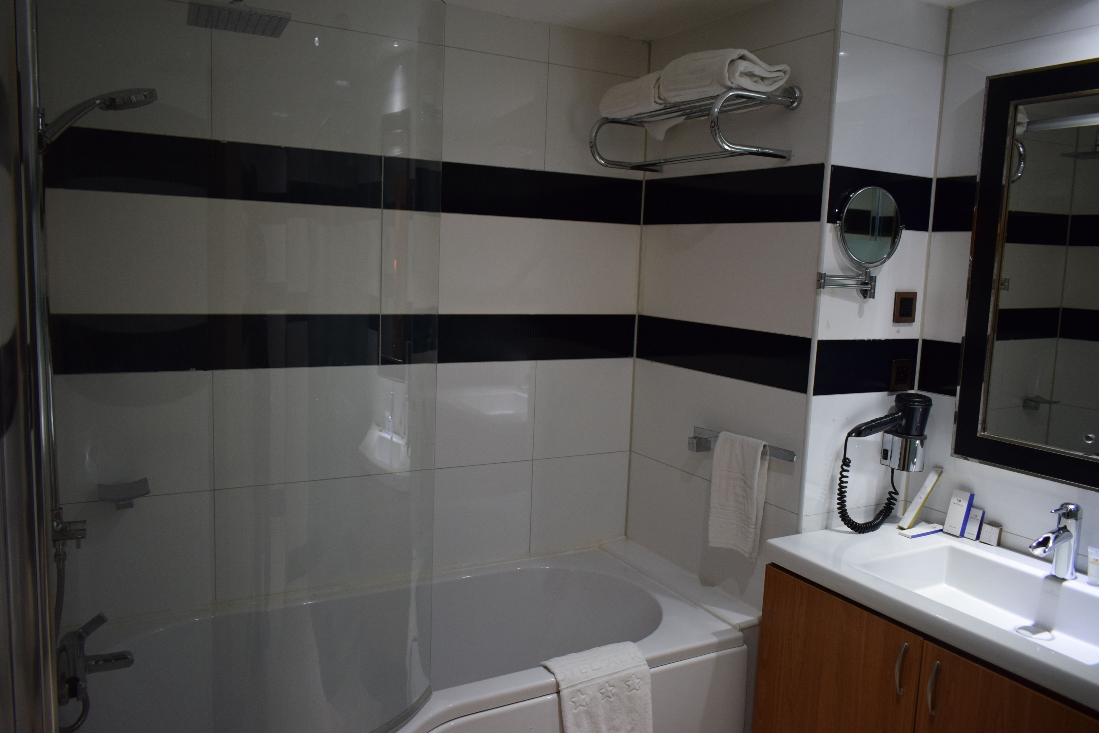 A bathroom with a glass-enclosed shower, a white bathtub, a wooden vanity with a mirror, and black and white striped tiles.