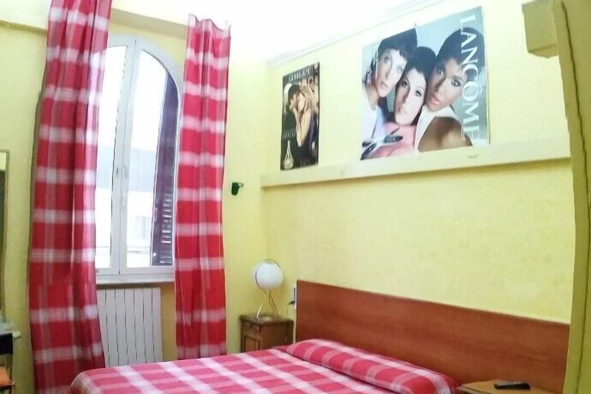 A bedroom with two beds, one with a checkered bedspread and the other with a floral bedspread. There is a window with red curtains, a nightstand with a lamp, and a poster on the wall.