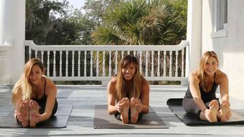 Three individuals performing a yoga pose on mats on a wooden deck with a white railing, surrounded by greenery.