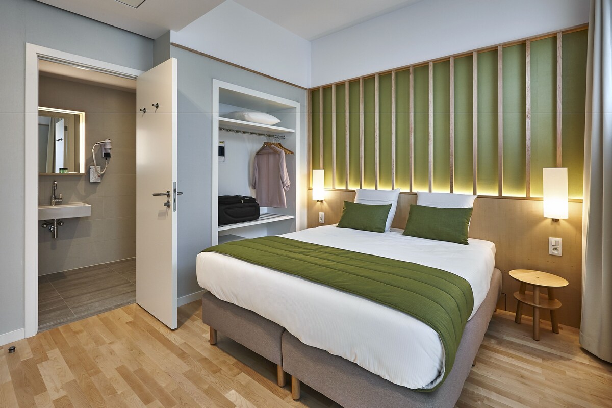 Una habitación de hotel con cama, mesita de noche, lámpara, armario y baño con espejo y lavamanos.
