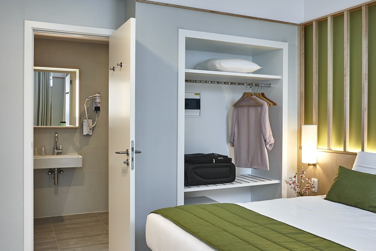 Une chambre d’hôtel comprenant un lit, un dressing, une salle de bain avec un miroir et une table de chevet avec une lampe.