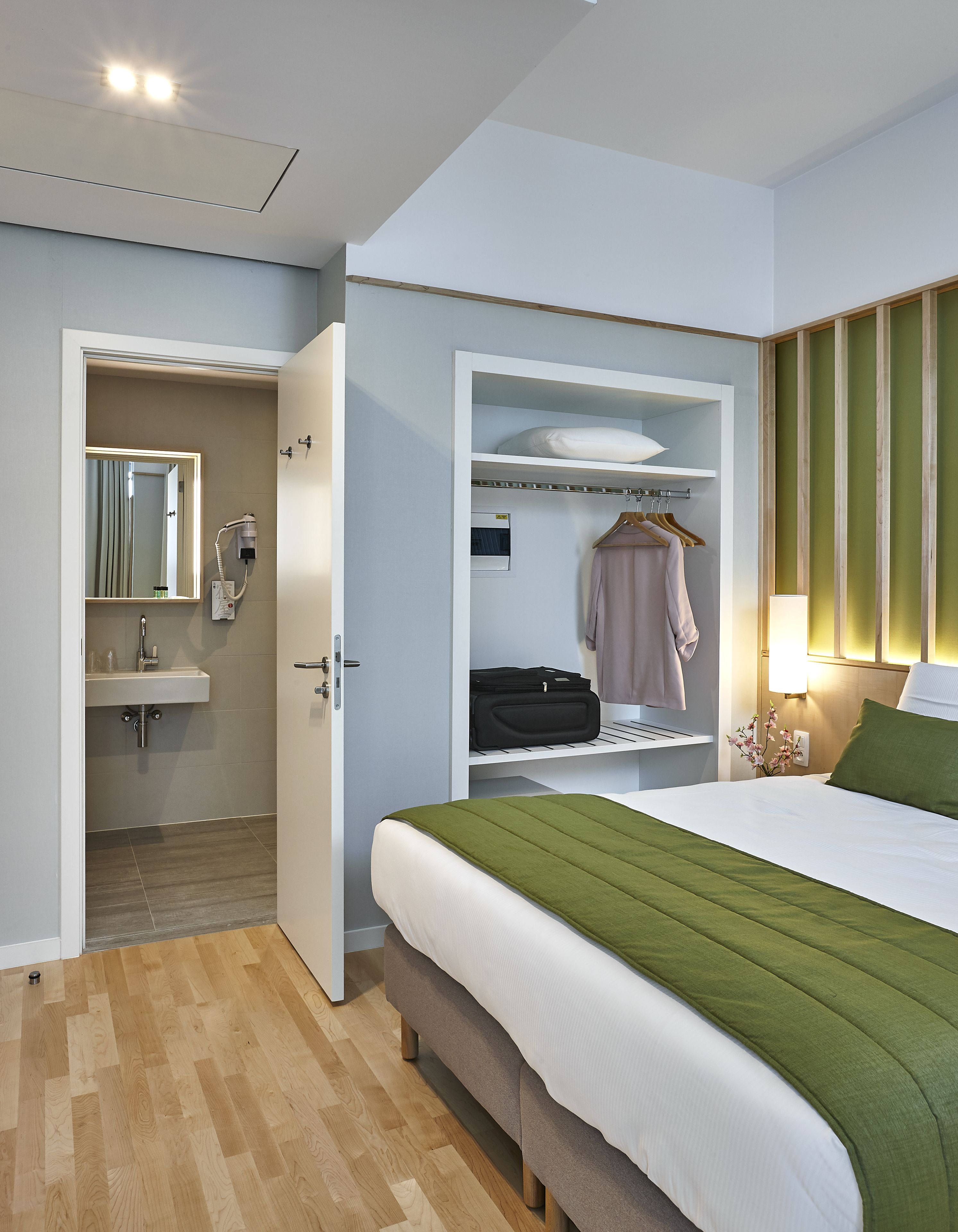 A hotel room with a bed, a walk-in closet, a bathroom with a mirror, and a bedside table with a lamp.