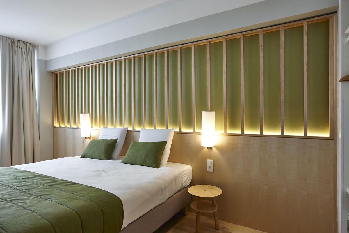 A hotel room with a bed, bedside lamps, a small round table, and a wooden headboard with vertical slats.