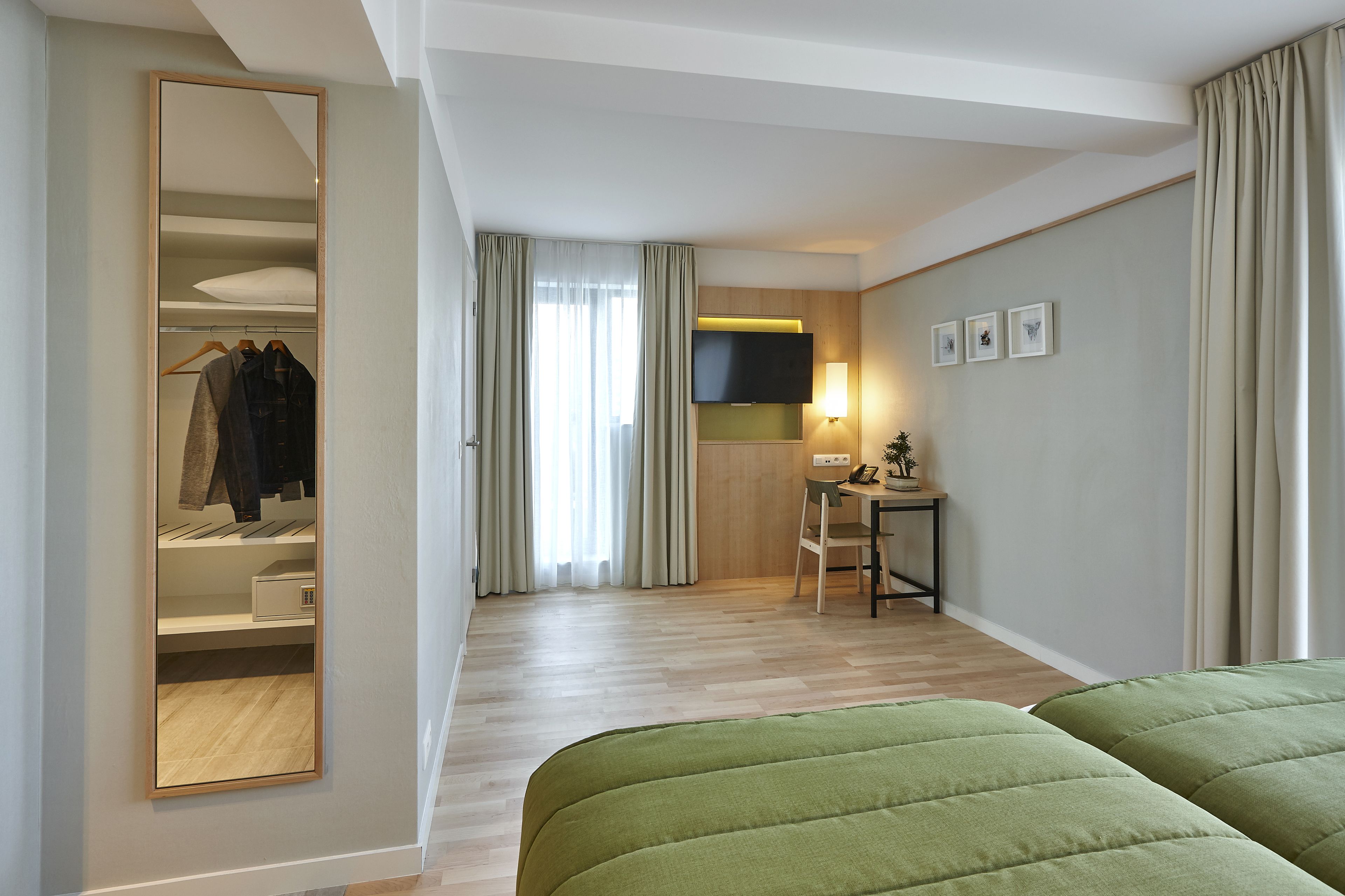 A modern hotel room with a large mirror, a desk with a chair, a flat-screen TV, and a bed with green bedding.
