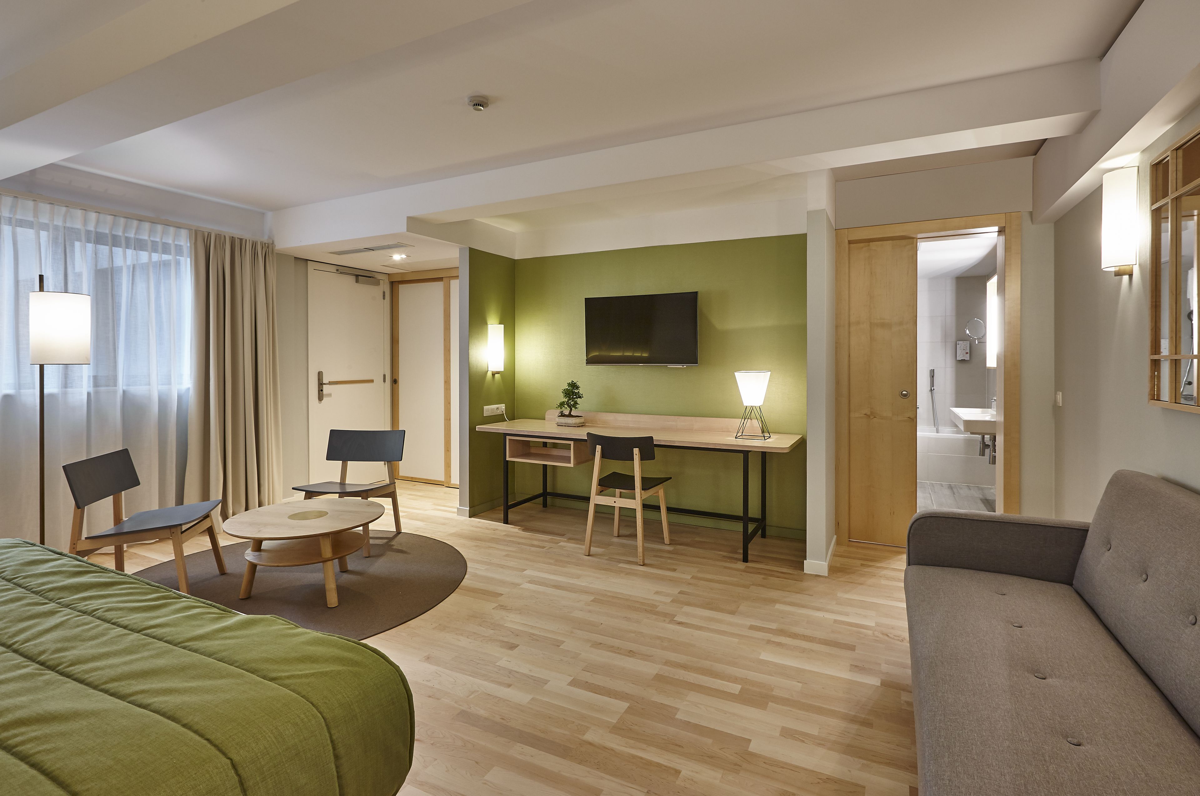 A hotel room with a bed, a sofa, a desk with a chair, a TV, and a bathroom visible through an open door.