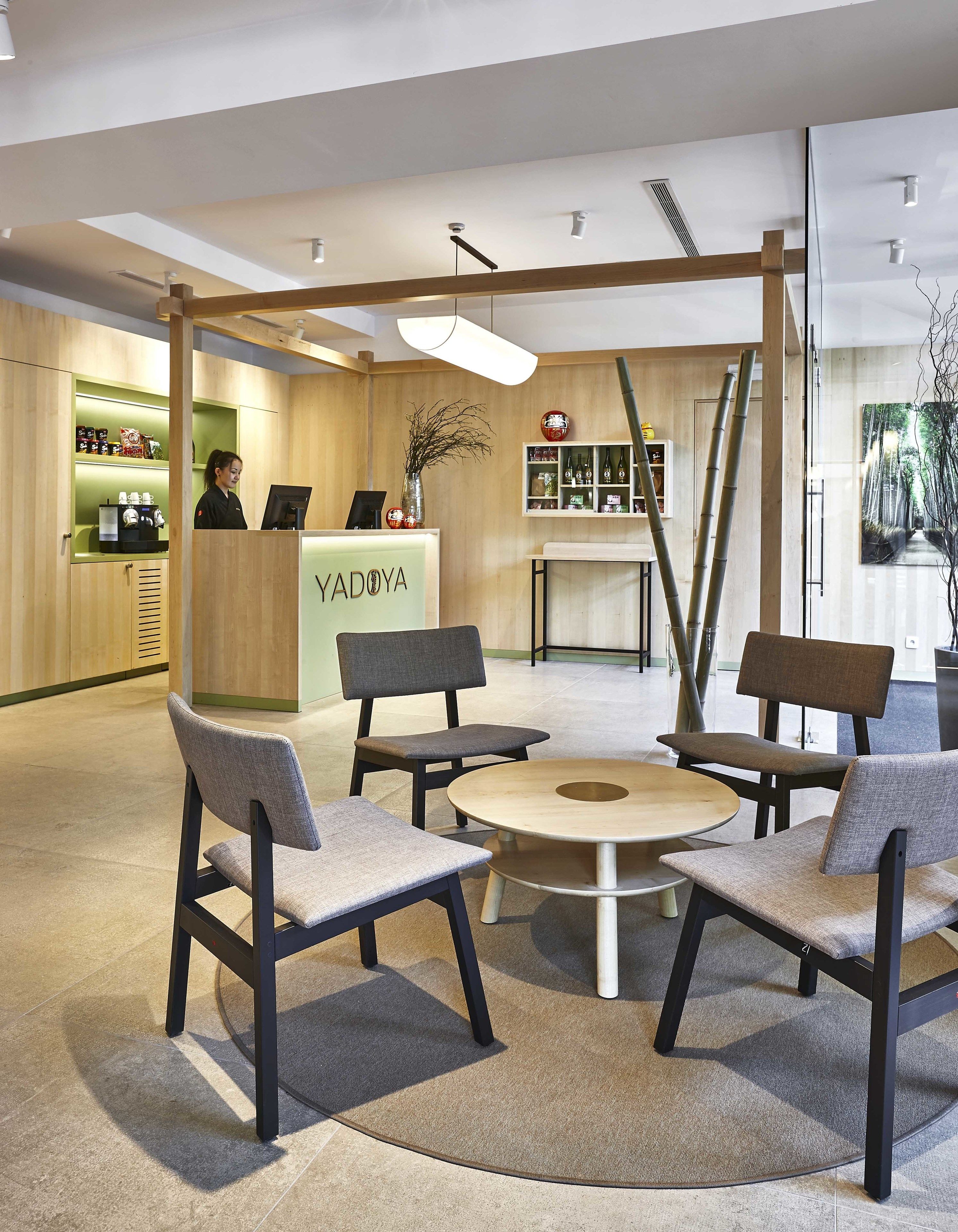 A modern interior with a reception desk labeled 'YADOYA', seating area with chairs and a round table, and a wooden wall with shelves and decorative items.