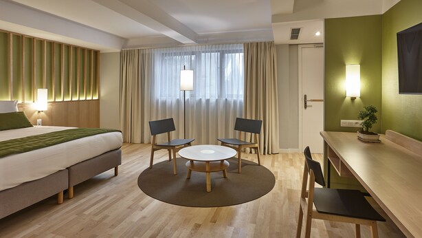 Une chambre d’hôtel avec un lit, une table ronde entourée de chaises, un bureau et une plante en pot.
