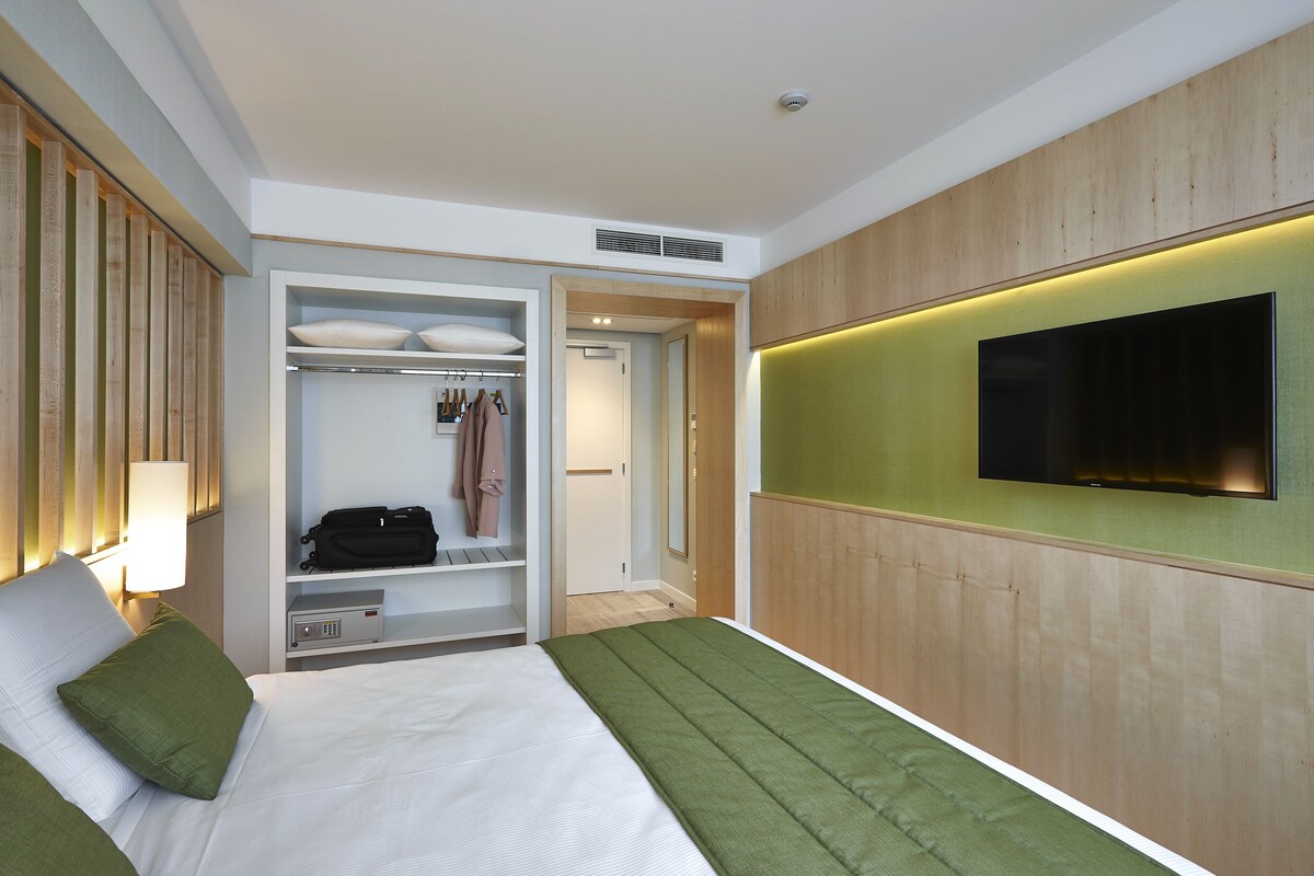 Habitación de hotel con cama, televisor montado en la pared, vestidor y mesita de noche con lámpara.