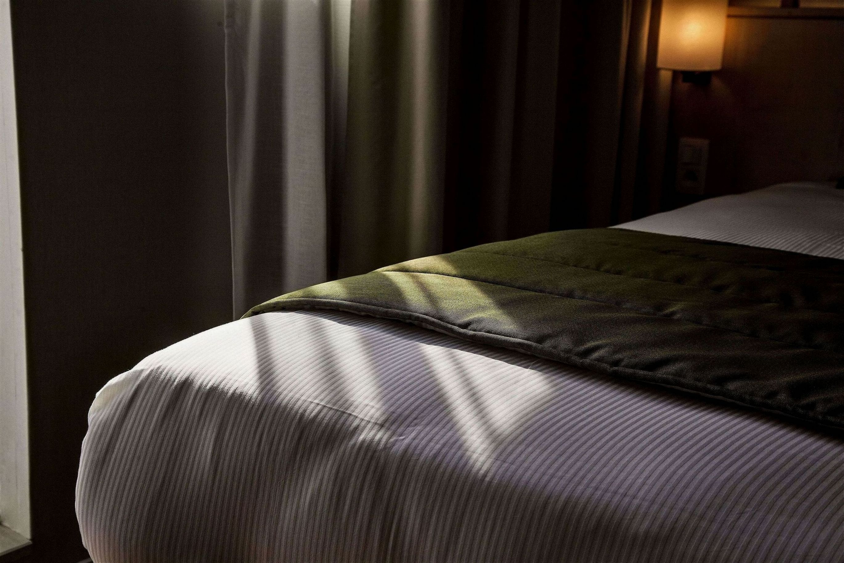 Un letto con una coperta verde, una lampada su un comodino e tende parzialmente aperte.