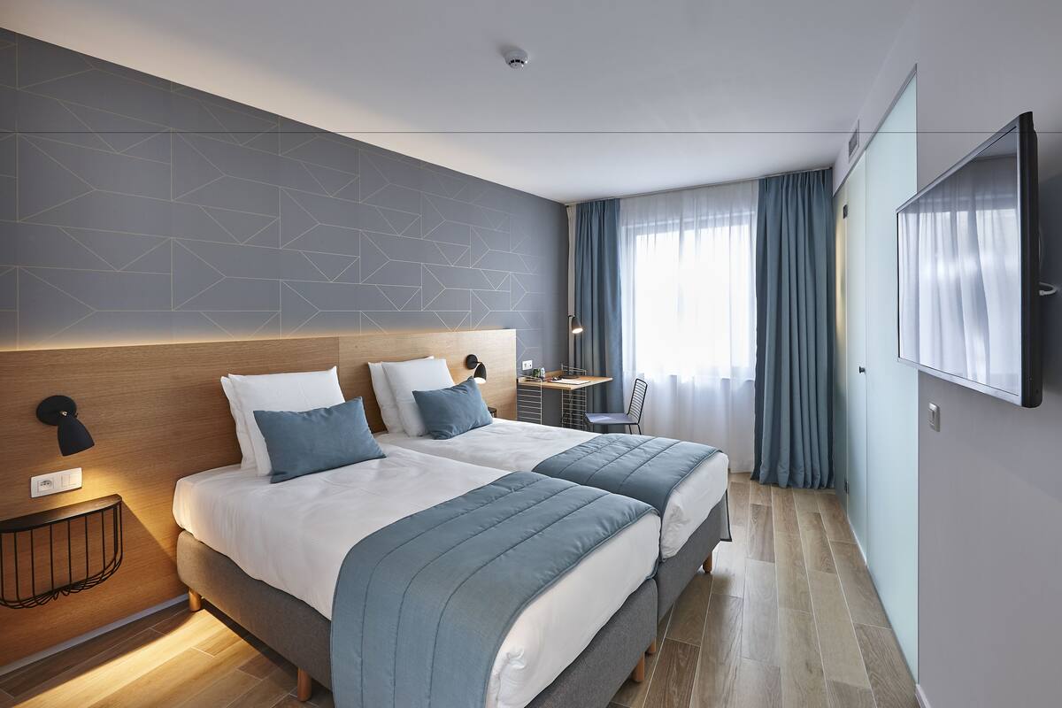 Ett modernt hotellrum med två sängar, ett skrivbord och ett stort fönster med gardiner.
