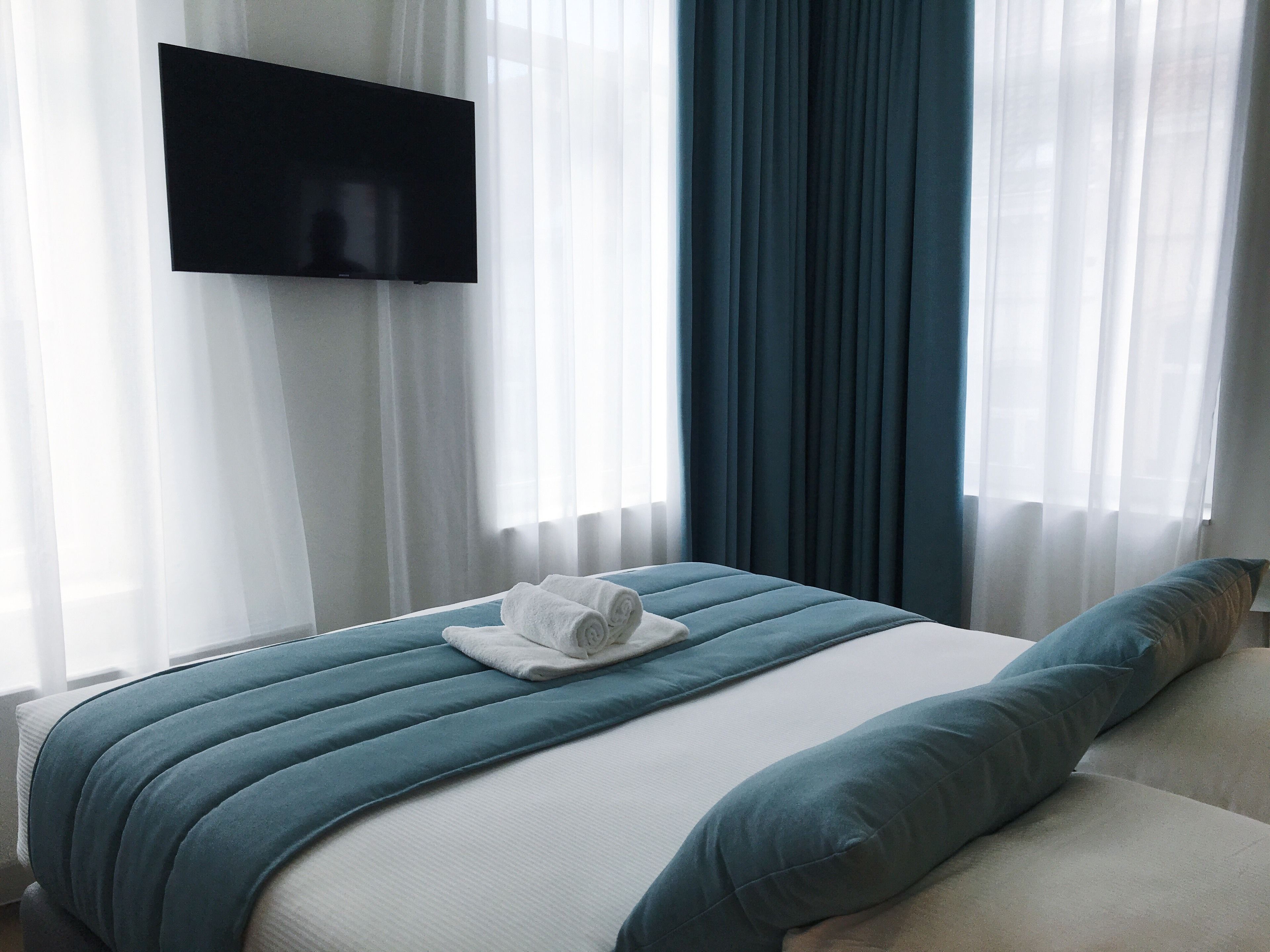 A hotel room with a bed, a flat-screen TV mounted on the wall, and sheer curtains.