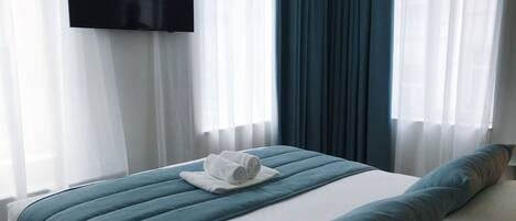 A hotel room with a bed, a flat-screen TV mounted on the wall, and sheer curtains.