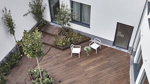 Un patio al aire libre con suelo de madera, sillas y mesas de metal blanco, rodeado de follaje y la fachada blanca de un edificio.