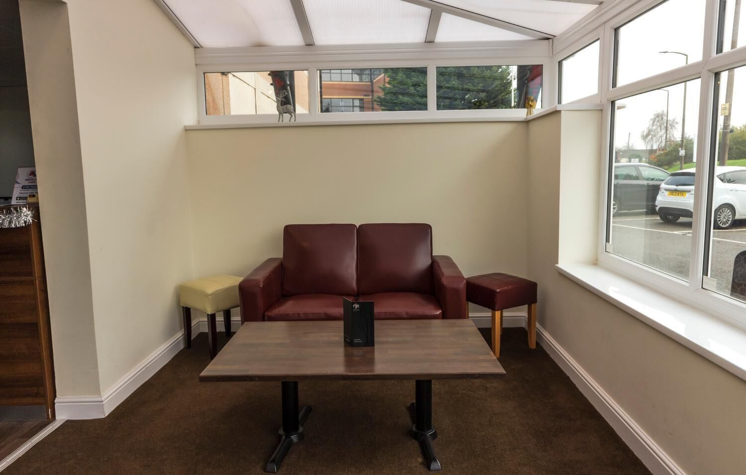 Una habitación con un sofá de cuero marrón, un pequeño taburete amarillo, una mesa de centro de madera y vistas a coches estacionados a través de amplias ventanas.