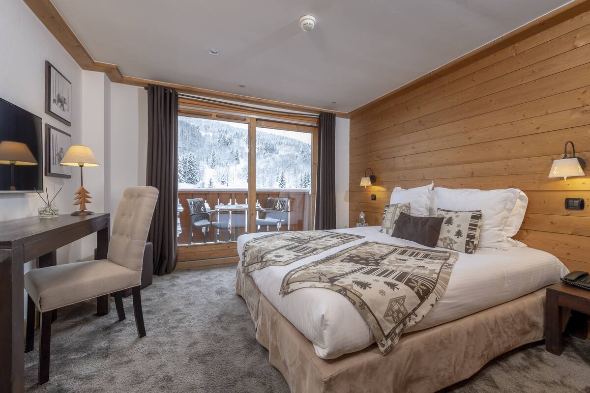 A bedroom with a large bed, a desk, and a chair. There is a window with a view of a snowy forest.
