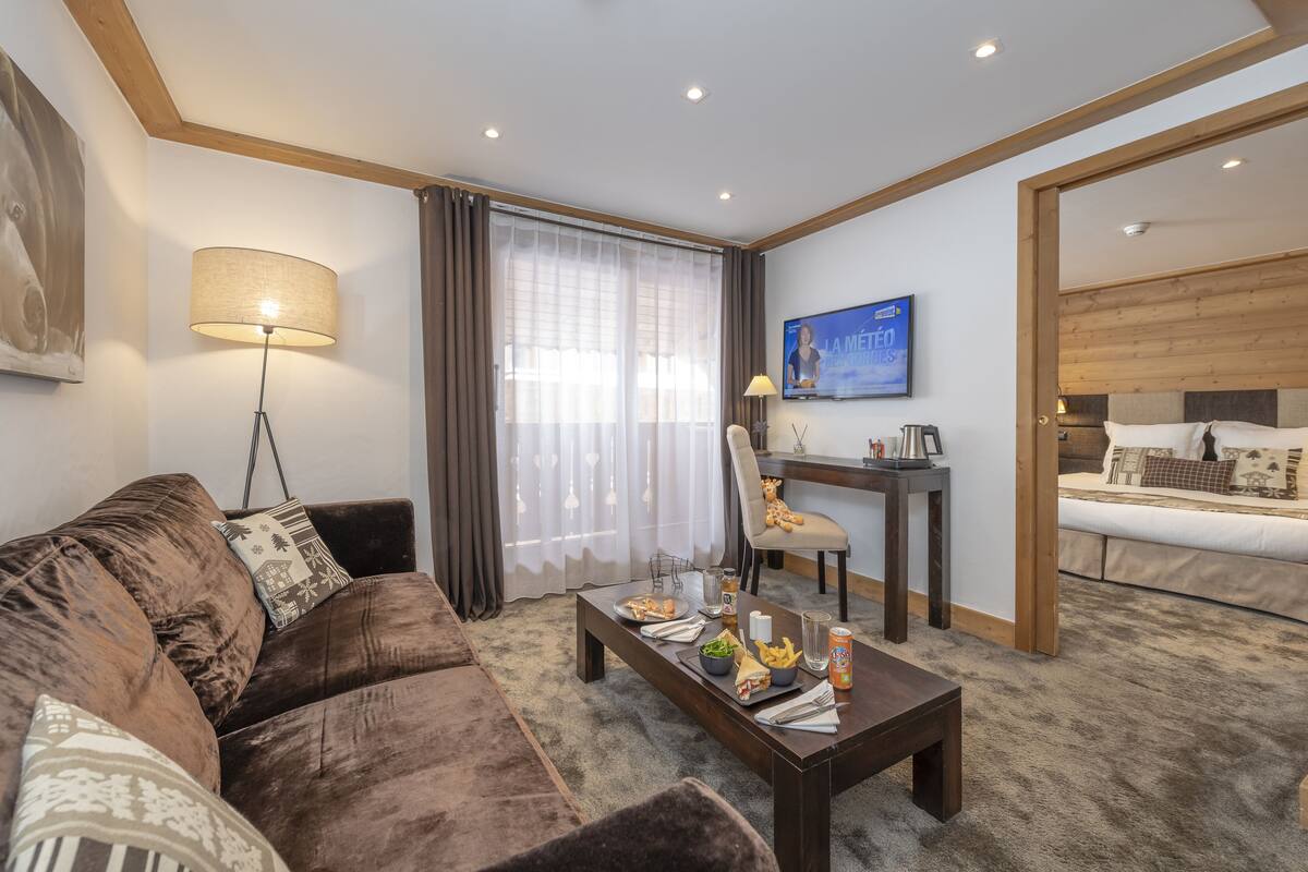 A modern hotel room with a brown sofa, a dining table with food, a TV, and a wooden bed.