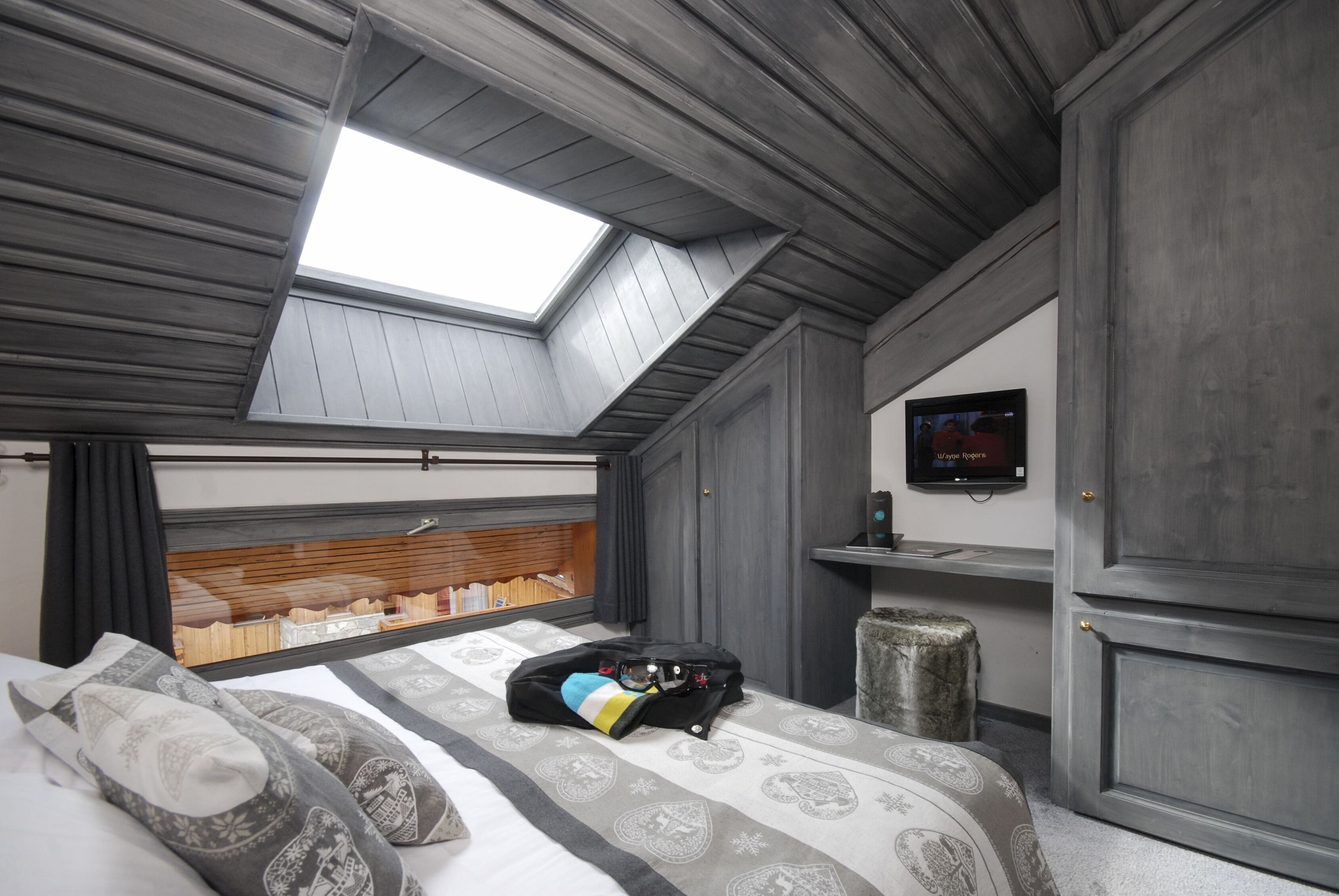 A bedroom with a skylight, a bed with patterned bedding, a suitcase, a television on the wall, and a wooden headboard.