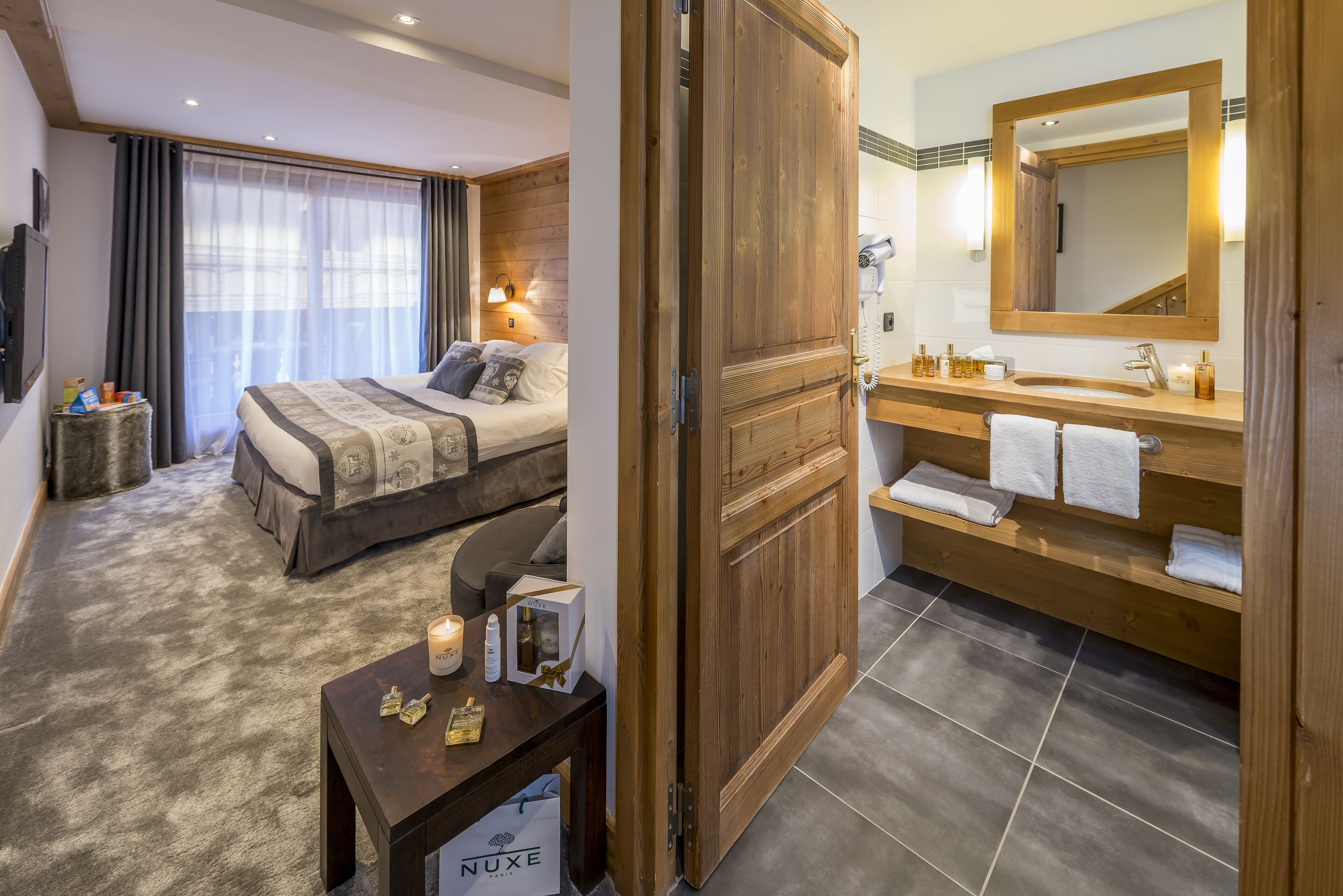 A hotel room with a bed, bedside table with toiletries, a wooden wardrobe, a bathroom with a sink and mirror, and a large window with curtains.