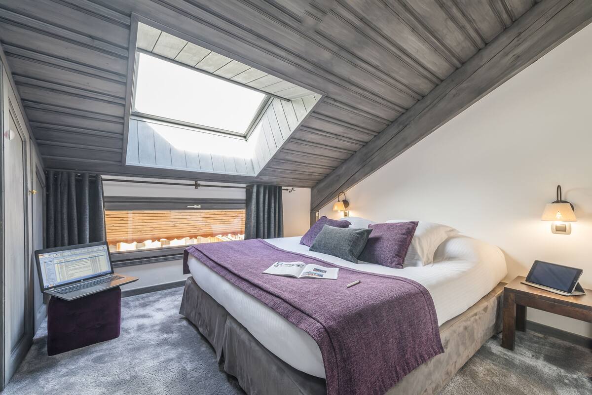 A bedroom with a skylight, a bed with a purple blanket, a laptop on a small table, and a bedside lamp.