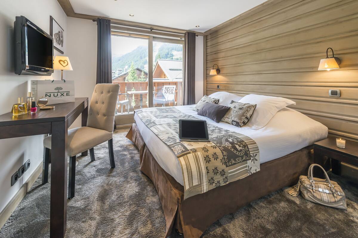 A hotel room with a bed, a desk with a laptop, a chair, a TV, and a view of mountains.
