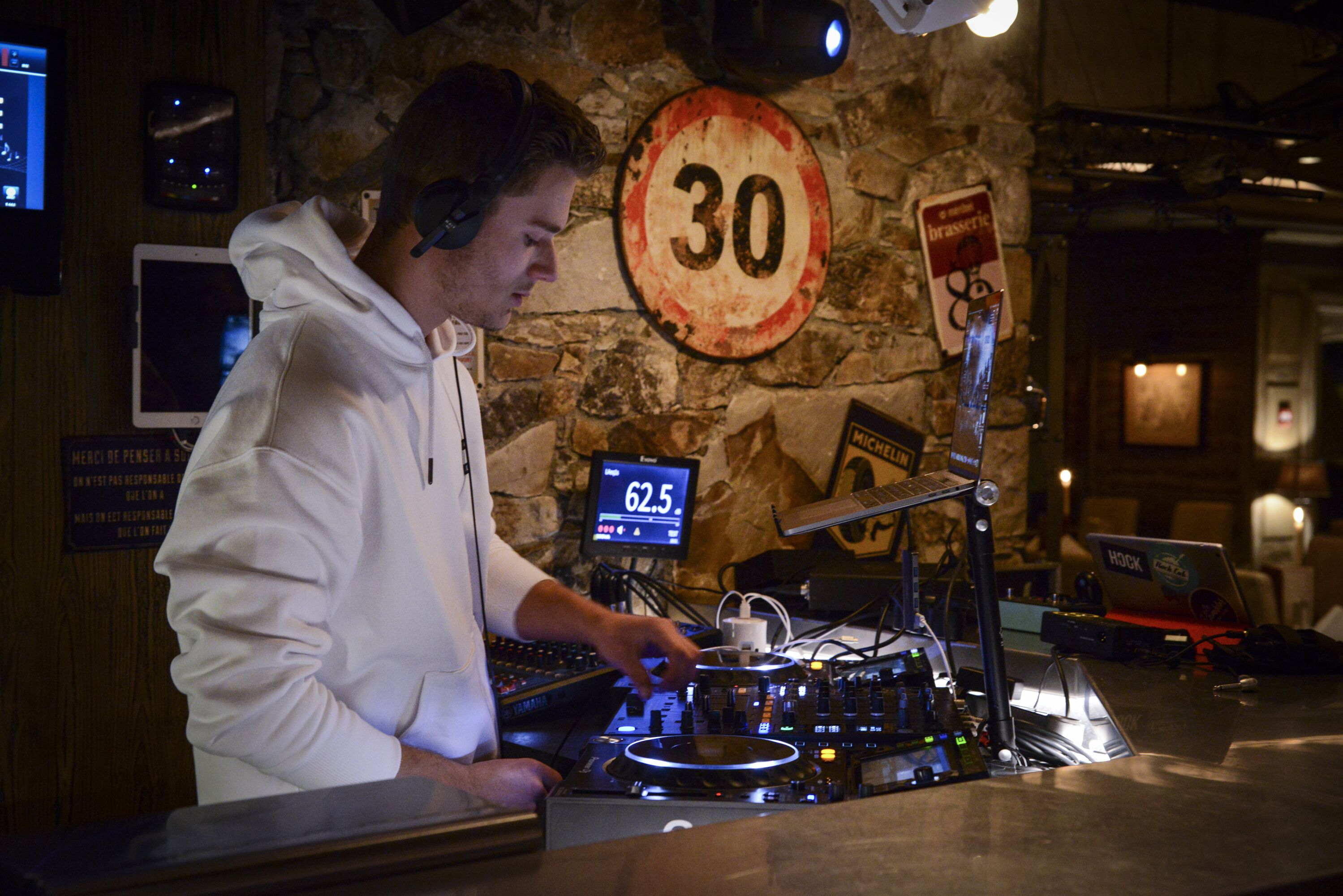Una persona manejando equipo de DJ en una habitación con paredes de piedra, una señal de límite de velocidad y una pantalla digital que muestra 62.5.