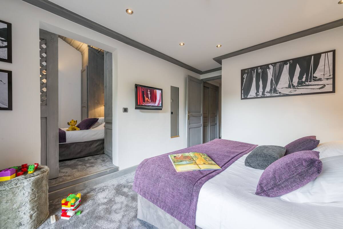 A bedroom with a bed, a purple blanket, and a book. There is a small table with toys and a mirror on the wall.
