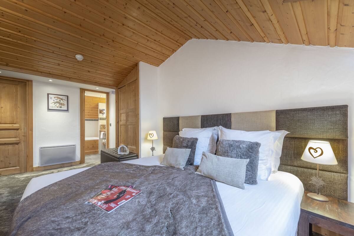A bedroom with a wooden ceiling, a large bed with multiple pillows, a nightstand with a lamp, and a wooden wardrobe.