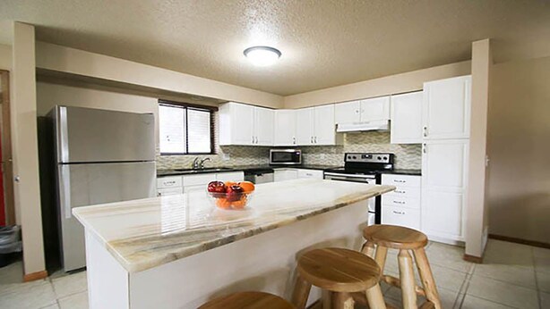 A modern kitchen with white cabinets, a marble countertop, a refrigerator, a microwave, a stove, and two wooden stools. - Woodside Dells Hotel & Suites (Wisconsin Dells)