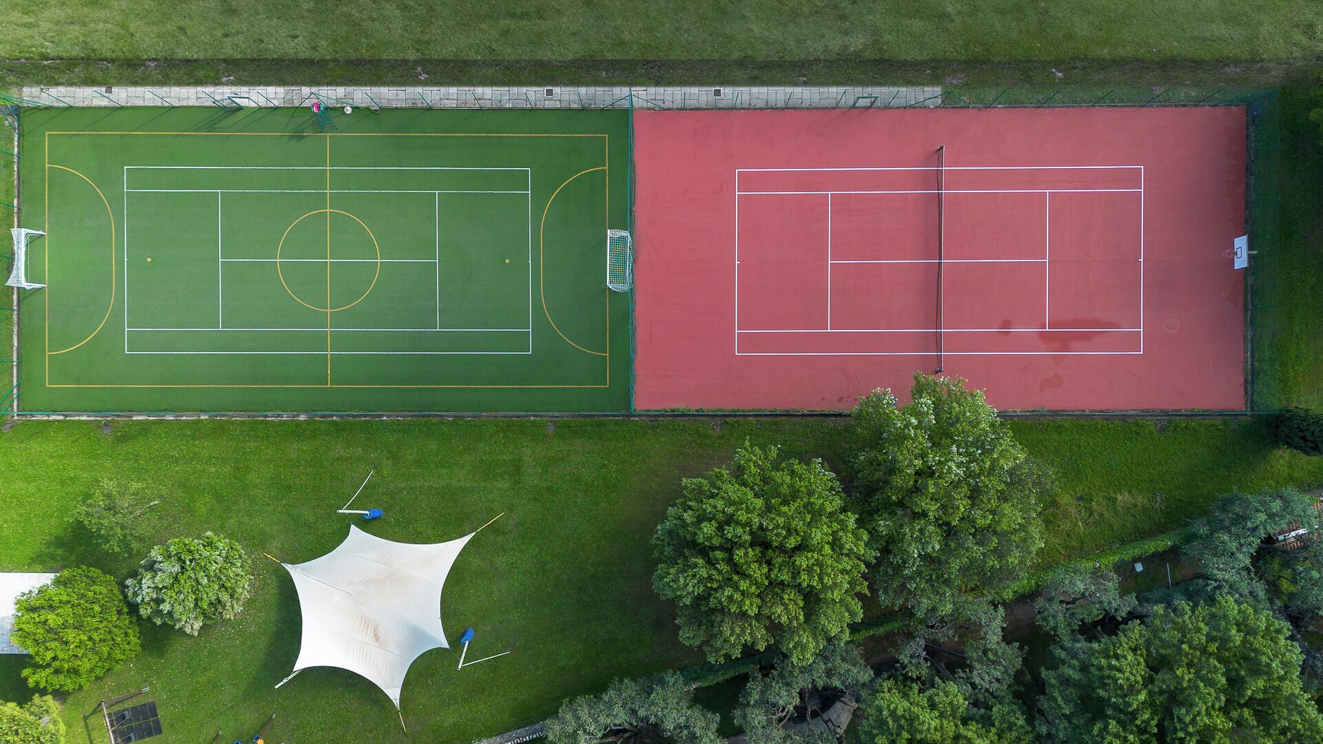 An aerial view of a sports facility with a soccer field and a tennis court, surrounded by trees and a white tent-like structure.