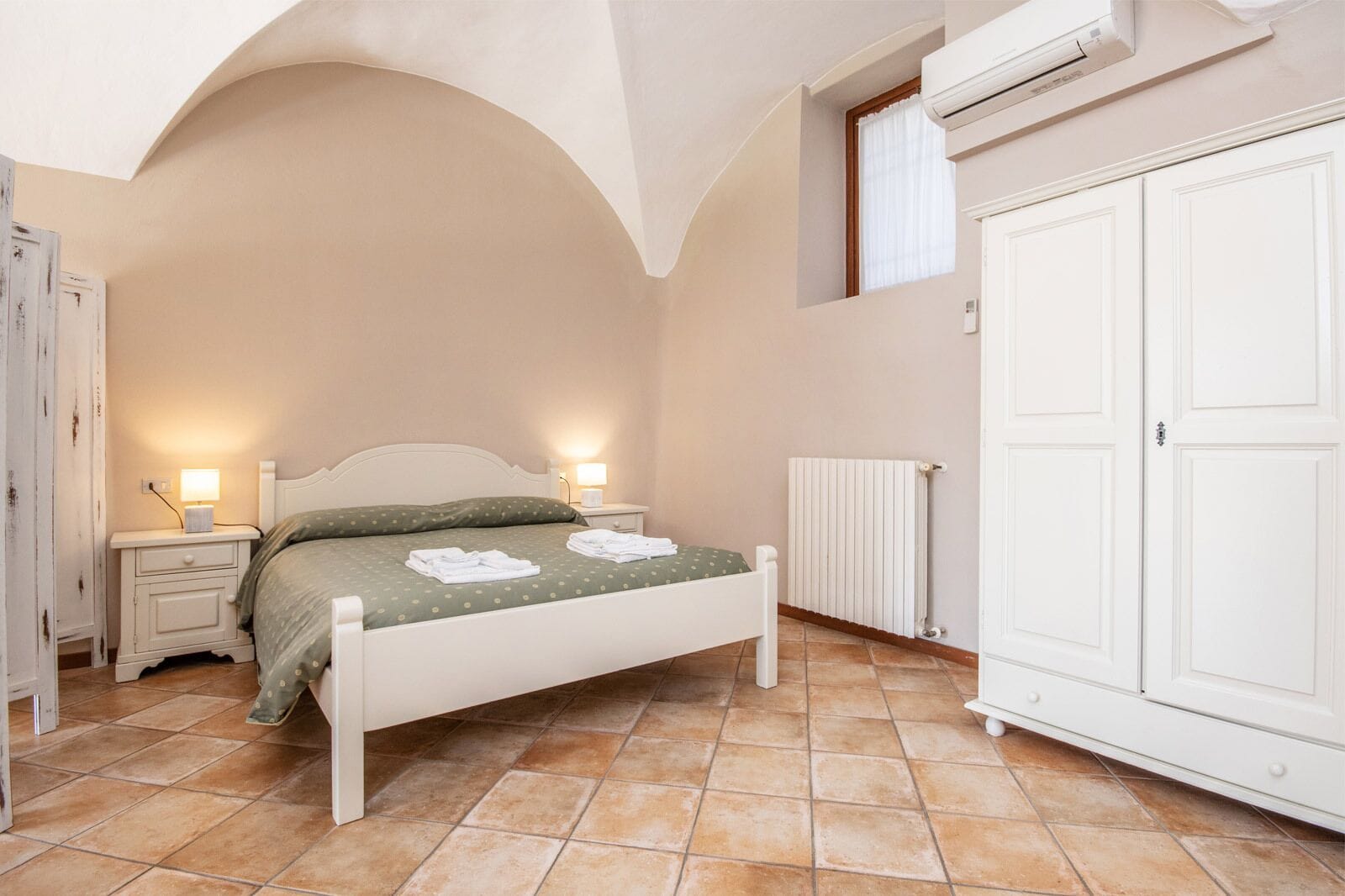 Un dormitorio con una cama, dos mesitas de noche con lámparas, un radiador y un armario.