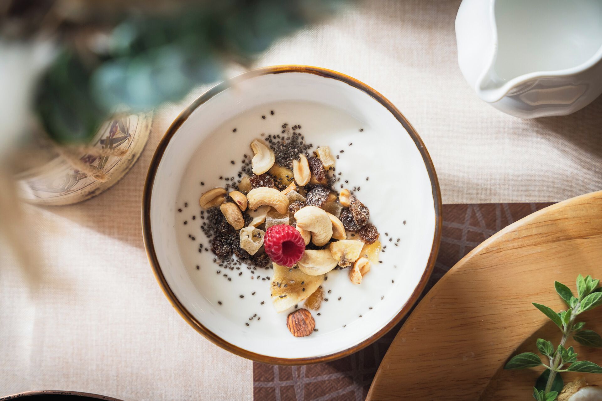 A bowl of yogurt topped with seeds, nuts, and a raspberry, placed on a table with a cup and a plant in the background.